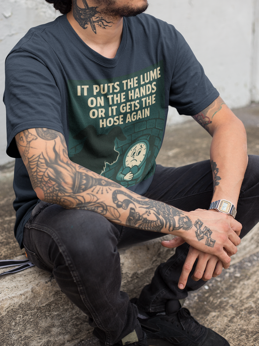 A man with tattoo covered arms wearing dark jeans sitting on a stone step, turned slightly to his left. He is wearing a pepper colored tshirt with a graphic in dark grayish green with a cream colored line drawing of an anamorphic watch face sitting at the bottom of a brick lined well with its knees drawn up to its chest, held by its arms. Looking down into the well is the silhouette of a small dog. The top of the graphic shows the text, in all caps, 'IT PUTS THE LUME ON THE HANDS OR IT GETS THE HOSE AGAIN'.