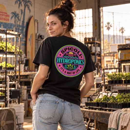 A woman wearing a black Spicoli Hydroponics graphic T-shirt, shown from the rear with a large retro surf-style back print in a vintage hydroponics shop setting.