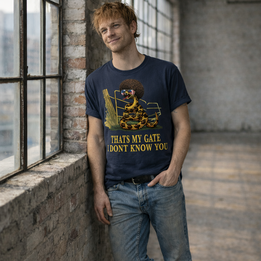 A man in a standing in a loft wearing a denim colored graphic t-shirt featuring a coiled rattlesnake, in front of a broken gate, with a large brown afro and flag-mirrored sunglasses above yellow text that reads "THAT'S MY GATE I DONT KNOW YOU."