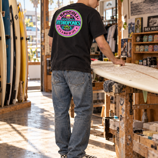 A young man in a gray knit beanie stands inside a SoCal surf shop, seen mostly from behind as he applies wax to a surfboard. He wears a black Spicoli Hydroponics T-shirt with a large colorful circular back graphic, worn blue jeans, and black-and-white checkerboard slip-on shoes. Surfboards, wax displays, posters, and warm natural light fill the shop around him.