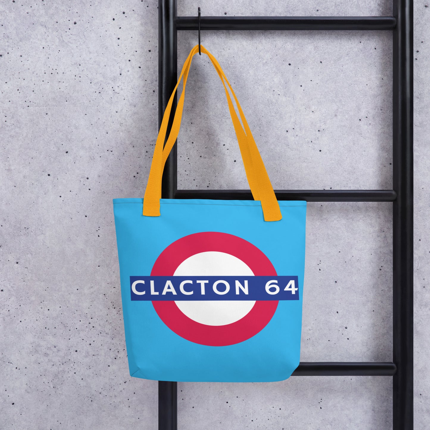 A tote bag with a light blue background and yellow straps. The graphic is a large red, white, and blue target-style roundel with a blue bar reading 'CLACTON 64'.