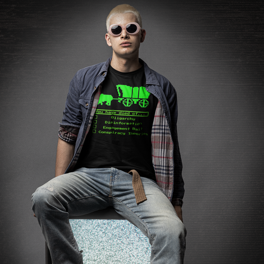 A man with cropped blonde hair wearing a black t-shirt with green text, sitting on a vintage television set against a dark background.