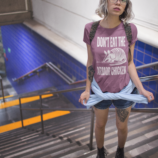 A woman wearing jean shorts with a tattooed left thigh, standing on a set of stairs, facing the top of the stairs and wearing a berry colored t-shirt with bold, all-caps, distressed white text that says 'DONT EAT THE ARMOR CHICKEN'. The text is separated into two lines by a white, line drawing graphic of an armadillo eating grass.