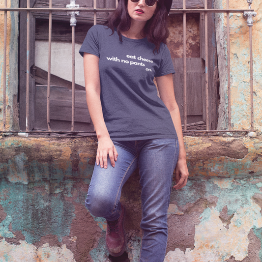 A woman wearing jeans, sunglasses and cherry Doc Martens leaning against a window sill with her right leg bent at the knee and her foot on the wall. She is wearing a midnight blue t-shirt with text that reads 'eat cheese with no pants on' on a gray background