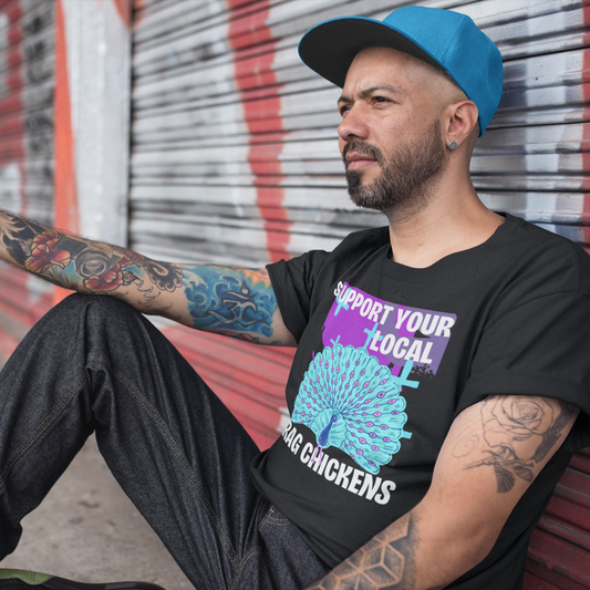 A man sitting on the ground leaning against a graffiti colored steel roll up door. He is wearing a black colored short-sleeve crewneck t-shirt featuring a stylized graphic of a peacock with its tail feathers fully fanned out. The peacock is light blue with pink eye details, set against a distressed purple background block with light blue plus signs. Above the peacock is bold white text reading 'SUPPORT YOUR LOCAL', and below it reads 'DRAG CHICKENS'.
