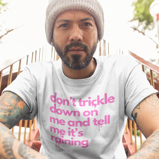 A bearded man sitting on a staircase leaning toward the viewer with his hands folded to the front. He has a full beard and is wearing a white colored t-shirt with pink text reading 'don't trickle down on me and tell me it's raining' in lower case letters with a dripping effect on some of the letters.