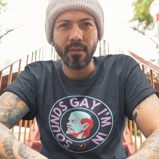 A man with a full beard & moustache, wearing a beige beanie, sitting on stairs with his hands crossed in front of himself but out of sight. He has heavily tattooed arms & is staring directly into the camera. He is wearing a true navy colored t-shirt. The chest shows a graphic of an art deco style bust of Lenin's right profile with a light blue face & red hair, beard & eyebrows surrounded by a line circle in the same light blue. An outer circle of bold all caps neon pink text reads 'SOUNDS GAY I'M IN'.