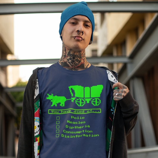 A man with facial piercings and tattoos covering his neck wearing a black jacket with a colorful lining. He is holding the jacket open with his left hand to reveal a navy blue short-sleeve crewneck t-shirt featuring a bright green, retro 8-bit pixel graphic. The design shows an ox pulling a covered wagon above a text box that reads 'You have died of...'. Below the text is a checklist with empty square boxes next to the words 'Polio', 'Measles', 'Diptheria', 'Consumption', and 'Disinformation'.