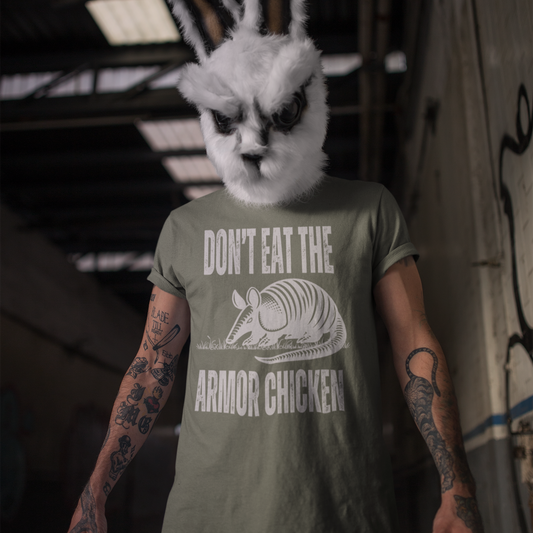 A man with heavily tattooed arm wearing a frightening rabbit head costume standing in a graffit covered hallway. He is wearing a moss green t-shirt with bold, all-caps, distressed white text that says 'DONT EAT THE ARMOR CHICKEN'. The text is separated into two lines by a white, line drawing graphic of an armadillo eating grass.