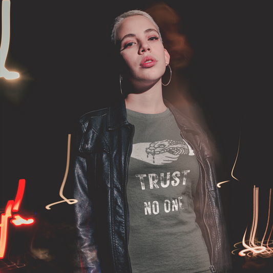 A woman with short blonde hair wearing a black leather jacket and a pepper colored short-sleeve crewneck t-shirt featuring a white graphic of a human hand shaking a snake, positioned above distressed white text that reads 'TRUST NO ONE'. The background is dark with a flash photography effect.