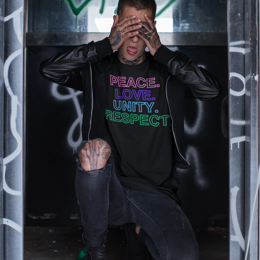 A man wearing a black leather jacket and black jeans kneeling on one knee and covering his face with tattooed hands. He is wearing black t-shirt with colorful text stacked in descending order that reads 'PEACE. LOVE. UNITY. RESPECT.'