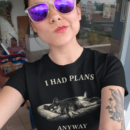 A woman wearing purple lensed sunglasses and making a 'duck face' with her lips toward the left side of her face. She is wearing a black colored t-shirt featuring a monochrome illustration of a long-haired Chihuahua sleeping deeply on its back in a dog bed, with rear legs splayed open and forepaws crossed. Above the illustration in distressed off-white capital letters is the text 'I HAD PLANS', and below it reads 'ANYWAY'.