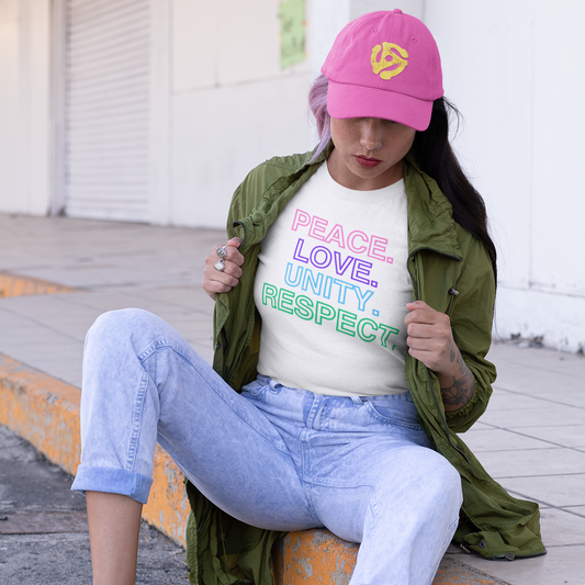 A woman sitting on a curb wearing a military green jacket that she is holding open at the chest to reveal a white t-shirt with colorful text stacked in descending order that reads 'PEACE. LOVE. UNITY. RESPECT.' on a gray background.