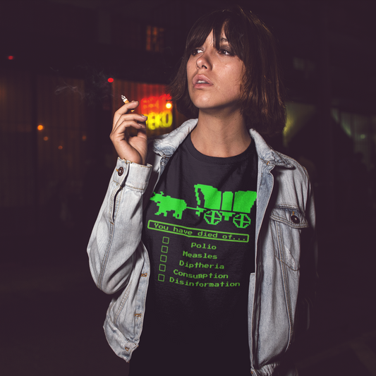 A woman wearing a jean jacket with her right arm raised just in front of her chin, holding a cigarette in her hand. She looks to her right while wearing a denim blue short-sleeve crewneck t-shirt featuring a bright green, retro 8-bit pixel graphic. The design shows an ox pulling a covered wagon above a text box that reads 'You have died of...'. Below the text is a checklist with empty square boxes next to the words 'Polio', 'Measles', 'Diptheria', 'Consumption', and 'Disinformation'