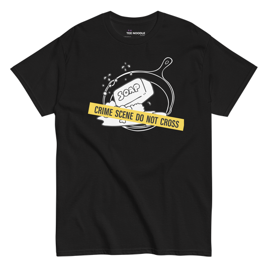 A black t-shirt featuring a white line-art graphic of a cast iron skillet filled with bubbles and a bar of soap. A yellow strip of tape across the front reads 'CRIME SCENE DO NOT CROSS'.