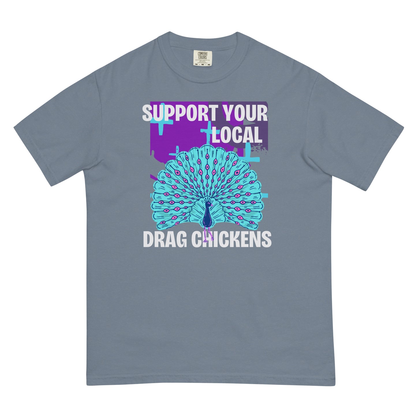 A blue jean colored short-sleeve crewneck t-shirt featuring a stylized graphic of a peacock with its tail feathers fully fanned out. The peacock is light blue with pink eye details, set against a distressed purple background block with light blue plus signs. Above the peacock is bold white text reading 'SUPPORT YOUR LOCAL', and below it reads 'DRAG CHICKENS'.