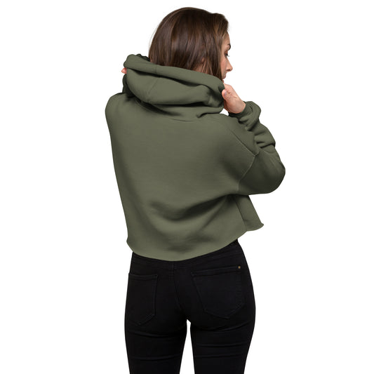 An athletic woman standing with her back to the viewer holding up the hood of a military green crop hoodie to display the length of hoodie at the waist.