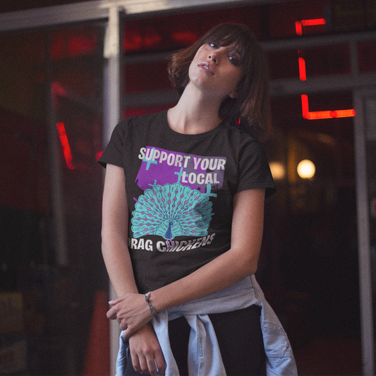A woman standing against an industrial, neon lit background. She is wearing a graphite colored short-sleeve crewneck t-shirt featuring a stylized graphic of a peacock with its tail feathers fully fanned out. The peacock is light blue with pink eye details, set against a distressed purple background block with light blue plus signs. Above the peacock is bold white text reading 'SUPPORT YOUR LOCAL', and below it reads 'DRAG CHICKENS'.