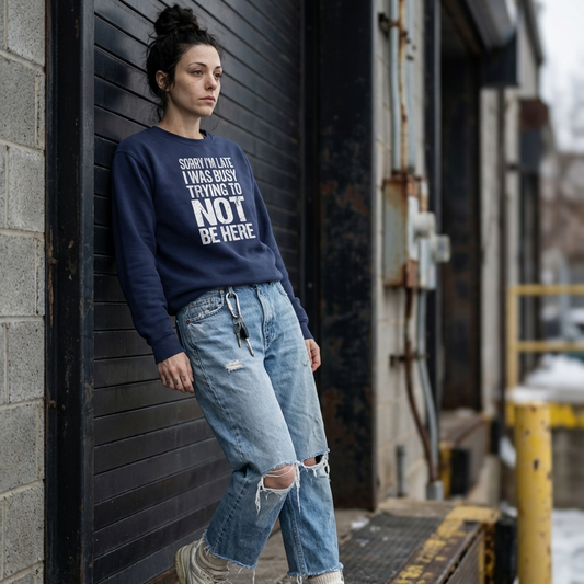A young woman with a messy top-knot leaning against a black industrial metal door, wearing an oversized navy blue crewneck sweatshirt that reads 'Sorry I'm Late I Was Busy Trying To Not Be Here' in bold white all-caps, distressed text. She is styled in baggy, distressed light-wash jeans and chunky sneakers.