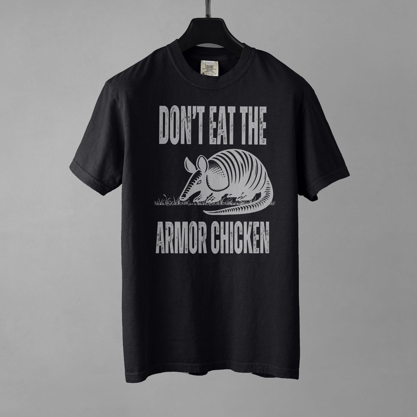 A black t-shirt with bold, all-caps, distressed white text that says 'DONT EAT THE ARMOR CHICKEN'. The text is separated into two lines by a white, line drawing graphic of an armadillo eating grass. 