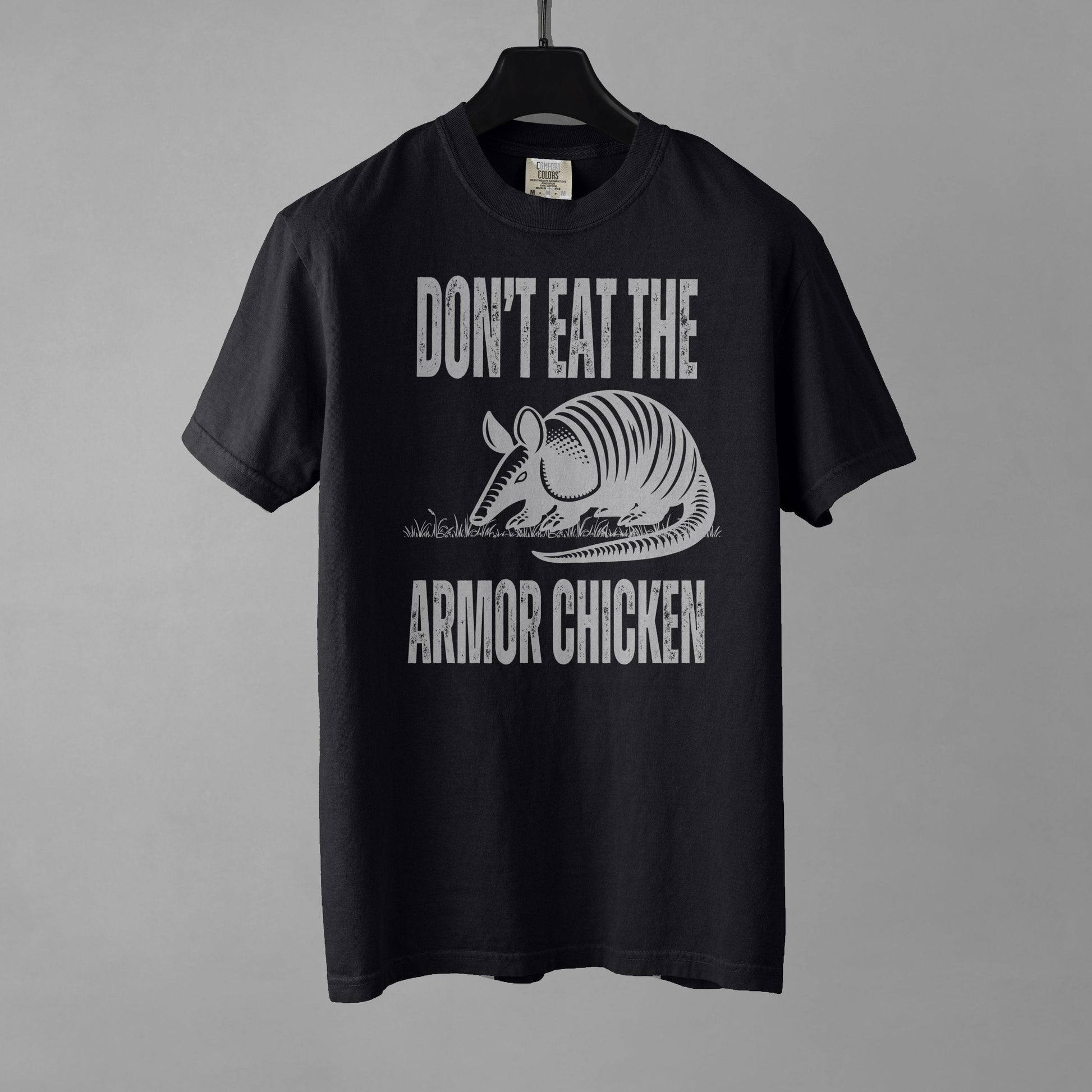 A black t-shirt with bold, all-caps, distressed white text that says 'DONT EAT THE ARMOR CHICKEN'. The text is separated into two lines by a white, line drawing graphic of an armadillo eating grass. 