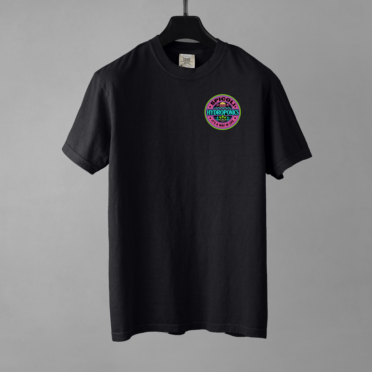 A black Comfort Colors 1717 T-shirt with a small Spicoli Hydroponics retro surf-style logo printed on the left chest. The garment is resting on a black hanger against a grey background.