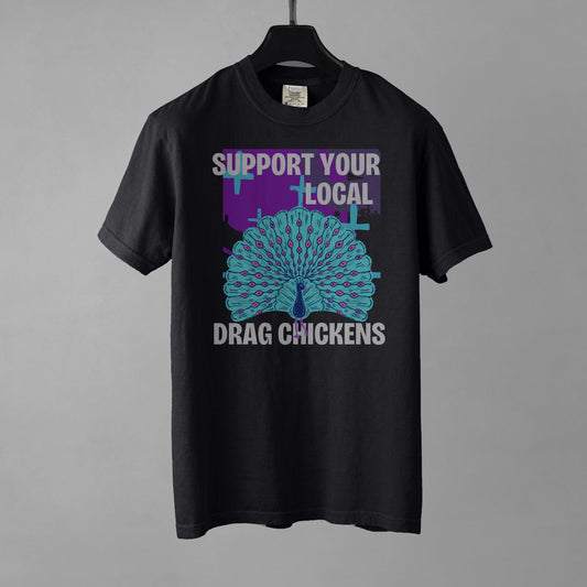 A black colored short-sleeve crewneck t-shirt featuring a stylized graphic of a peacock with its tail feathers fully fanned out. The peacock is light blue with pink eye details, set against a distressed purple background block with light blue plus signs. Above the peacock is bold white text reading 'SUPPORT YOUR LOCAL', and below it reads 'DRAG CHICKENS'. The shirt is presented on a black hanger against a gray background.