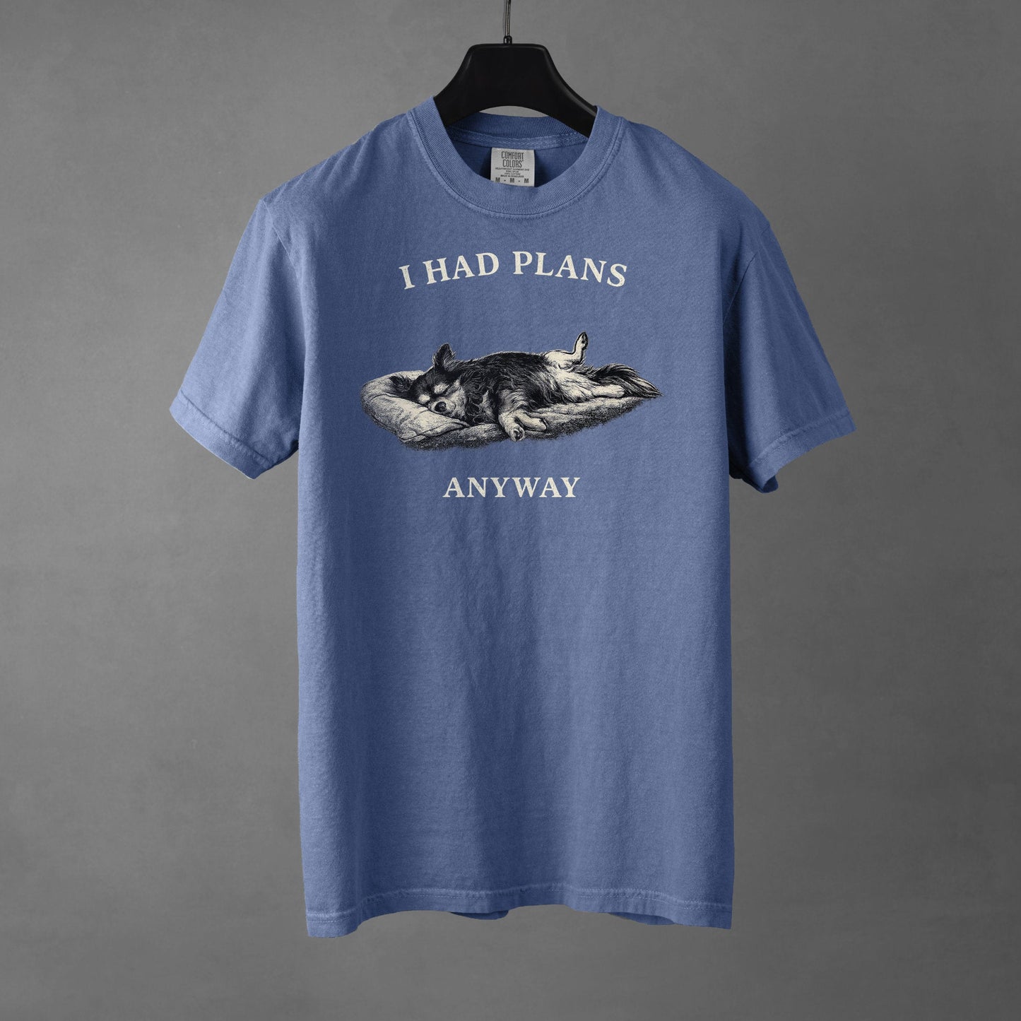 A blue jean colored t-shirt on a black hanger with a black and white graphic on the chest showing a long haired, tri-color Chihuahua laying its side on a dog bed with its rear legs splayed open and forepaws crossed. The Chihuahua is sleeping comfortably. Over the top of the graphic, in bold, white all-caps text is 'I HAD PLANS' while underneath it reads "ANYWAY', also in all-caps white text. 