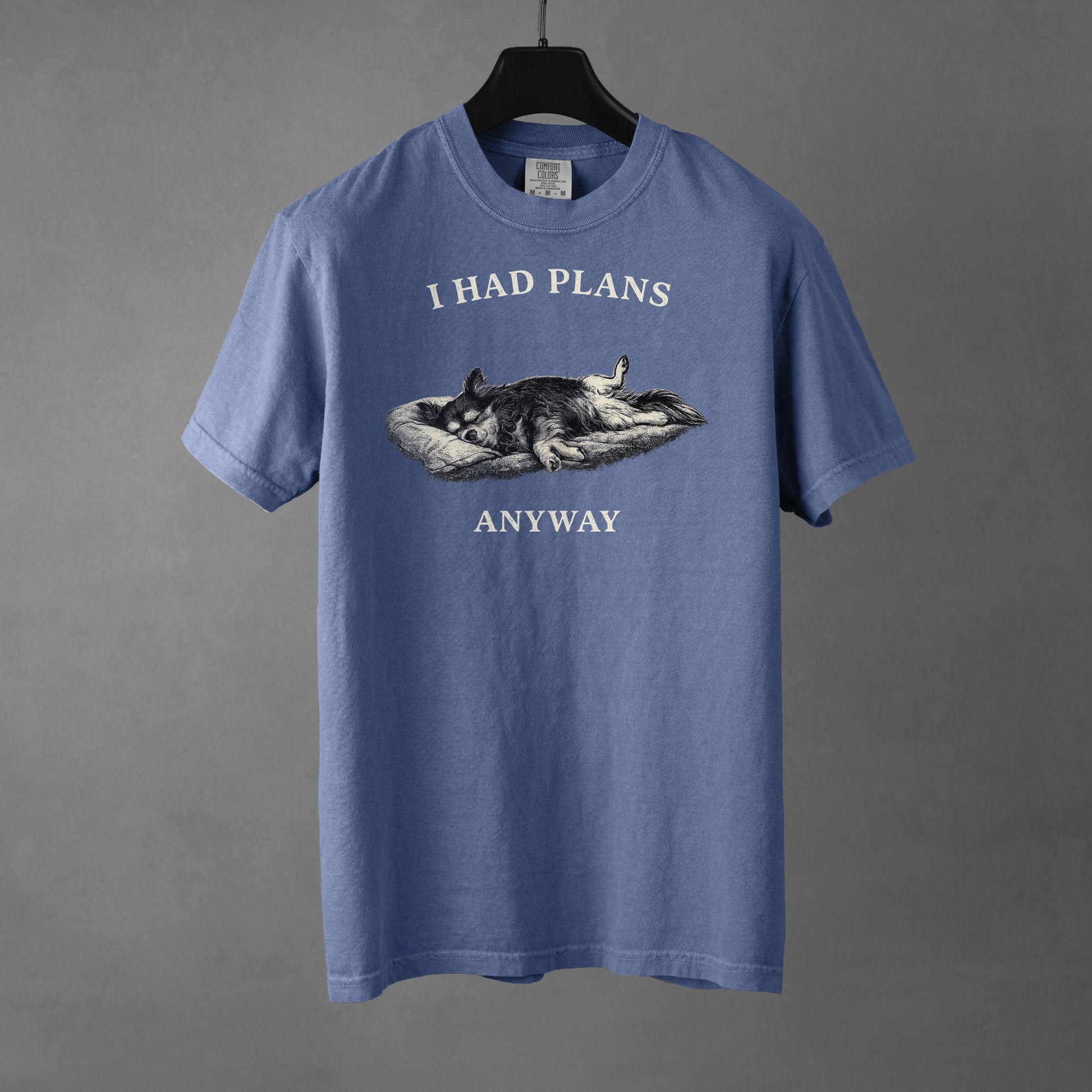 A blue jean colored t-shirt on a black hanger with a black and white graphic on the chest showing a long haired, tri-color Chihuahua laying its side on a dog bed with its rear legs splayed open and forepaws crossed. The Chihuahua is sleeping comfortably. Over the top of the graphic, in bold, white all-caps text is 'I HAD PLANS' while underneath it reads "ANYWAY', also in all-caps white text. 