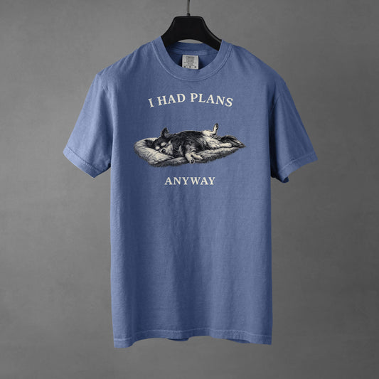 A blue jean colored t-shirt on a black hanger with a black and white graphic on the chest showing a long haired, tri-color Chihuahua laying its side on a dog bed with its rear legs splayed open and forepaws crossed. The Chihuahua is sleeping comfortably. Over the top of the graphic, in bold, white all-caps text is 'I HAD PLANS' while underneath it reads "ANYWAY', also in all-caps white text. 