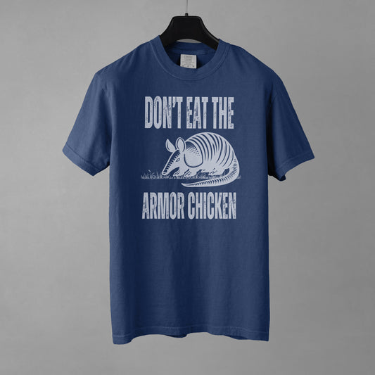 A denim colored t-shirt with bold, all-caps, distressed white text that says 'DONT EAT THE ARMOR CHICKEN'. The text is separated into two lines by a white, line drawing graphic of an armadillo eating grass. 