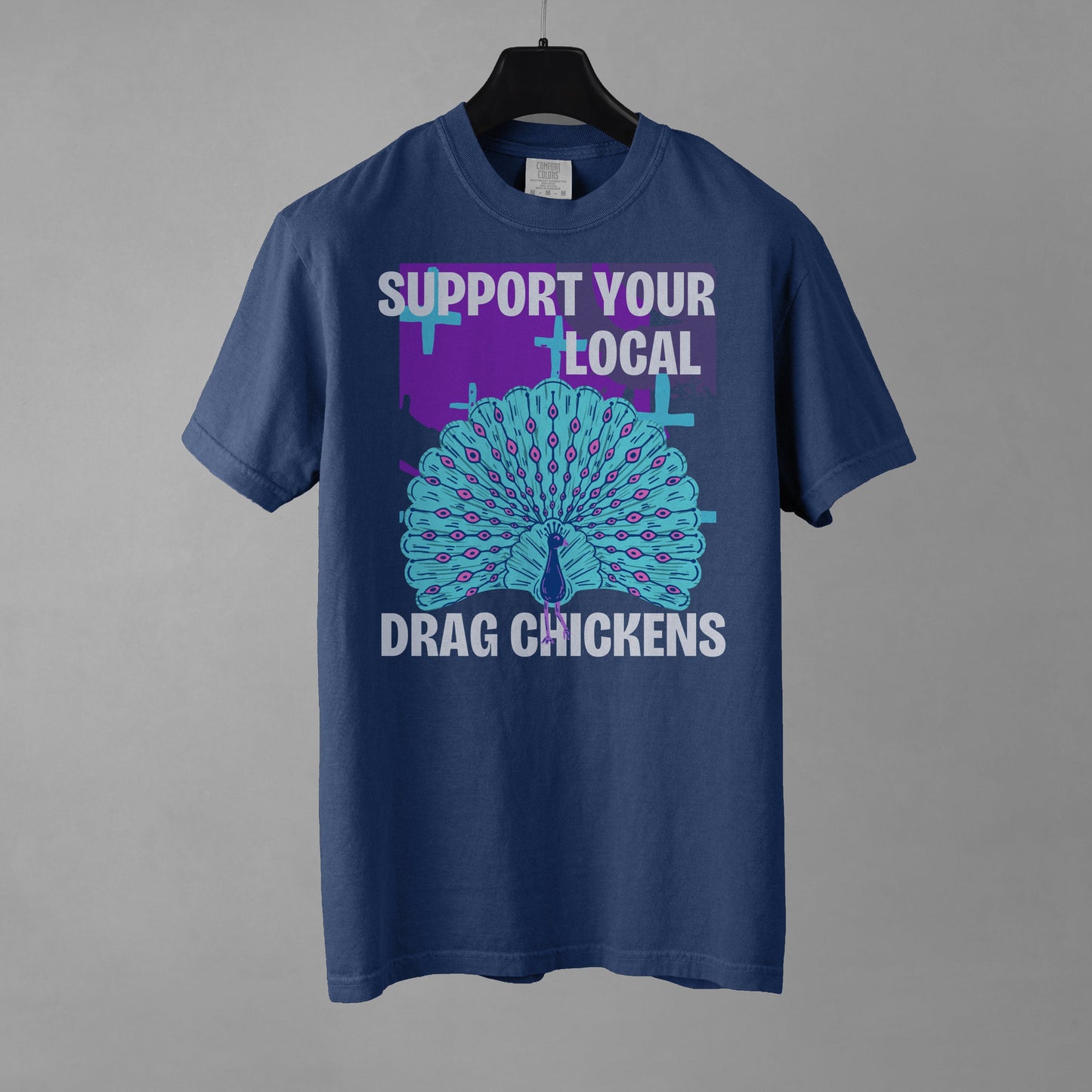 A denim colored short-sleeve crewneck t-shirt featuring a stylized graphic of a peacock with its tail feathers fully fanned out. The peacock is light blue with pink eye details, set against a distressed purple background block with light blue plus signs. Above the peacock is bold white text reading 'SUPPORT YOUR LOCAL', and below it reads 'DRAG CHICKENS'. The shirt is presented on a black hanger against a gray background.