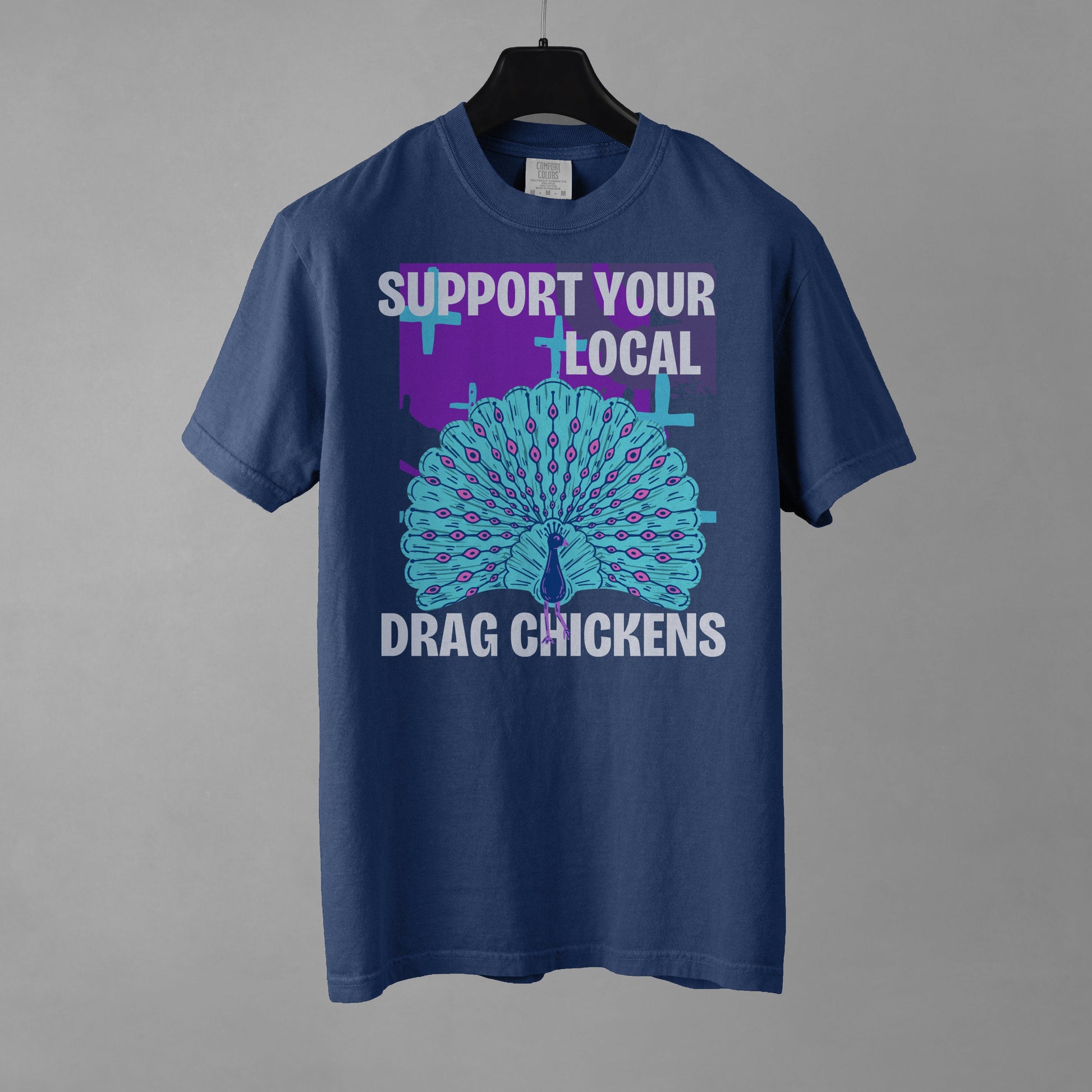 A denim colored short-sleeve crewneck t-shirt featuring a stylized graphic of a peacock with its tail feathers fully fanned out. The peacock is light blue with pink eye details, set against a distressed purple background block with light blue plus signs. Above the peacock is bold white text reading 'SUPPORT YOUR LOCAL', and below it reads 'DRAG CHICKENS'. The shirt is presented on a black hanger against a gray background.