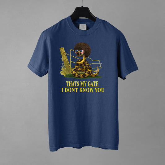 A denim colored t-shirt featuring a coiled rattlesnake with a large afro and flag sunglasses standing next to a farm gate with the text "THATS MY GATE I DONT KNOW YOU." The shirt is resting on a black hanger against a gray background.