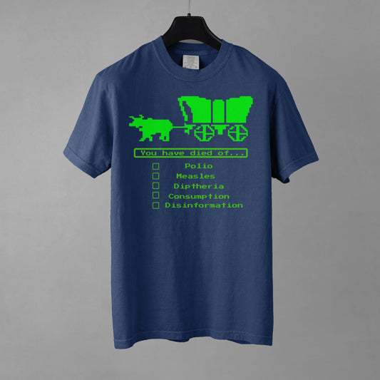 A denim blue short-sleeve crewneck t-shirt featuring a bright green, retro 8-bit pixel graphic. The design shows an ox pulling a covered wagon above a text box that reads 'You have died of...'. Below the text is a checklist with empty square boxes next to the words 'Polio', 'Measles', 'Diptheria', 'Consumption', and 'Disinformation'