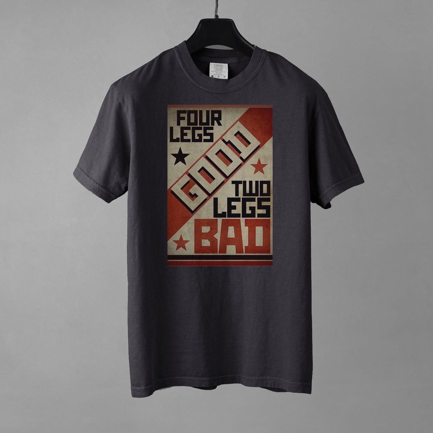 A graphite colored short-sleeve crewneck t-shirt featuring a retro propaganda-style rectangular graphic. The design uses a textured tan, red, and black color palette with diagonal geometric blocking. It features the text 'FOUR LEGS' in black, a large diagonal 'GOOD' in off-white, 'TWO LEGS' in black, and a large 'BAD' in red at the bottom. The design is accented with small black and red stars. The shirt is placed on a black hanger against a gray background.