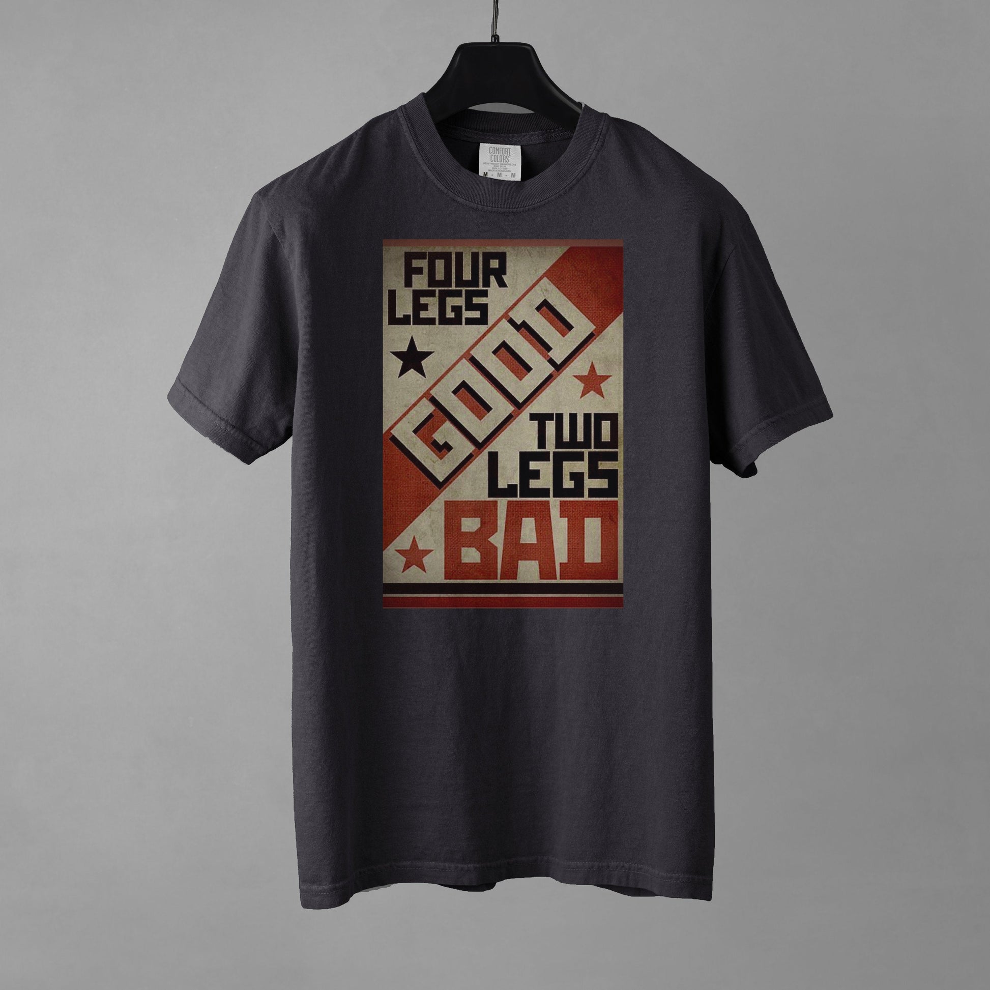 A graphite colored short-sleeve crewneck t-shirt featuring a retro propaganda-style rectangular graphic. The design uses a textured tan, red, and black color palette with diagonal geometric blocking. It features the text 'FOUR LEGS' in black, a large diagonal 'GOOD' in off-white, 'TWO LEGS' in black, and a large 'BAD' in red at the bottom. The design is accented with small black and red stars. The shirt is placed on a black hanger against a gray background.