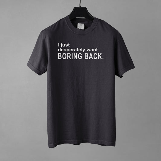 A graphite colored graphic t-shirt with the words "I just desperately want BORING BACK." printed in bold white sans-serif text across the chest. The shirt is resting on a black hanger against a gray background.