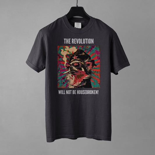 A graphite colored crewneck t-shirt featuring a vibrant, pop-art style square graphic on the chest. The graphic shows a long-haired Chihuahua stylized as a revolutionary, wearing a black hat with a red star, red-tinted sunglasses, and smoking a cigarette, set against a colorful, grunge-textured teal and red sunburst background. Above the graphic in white all-caps is the text 'THE REVOLUTION', and below it reads 'WILL NOT BE HOUSEBROKEN!'. The shirt is placed on a black hanger against a gray background.