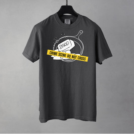 A pepper t-shirt with a graphic of a cast iron pan with a bar of soap in it, crossed with crime scene tape.
