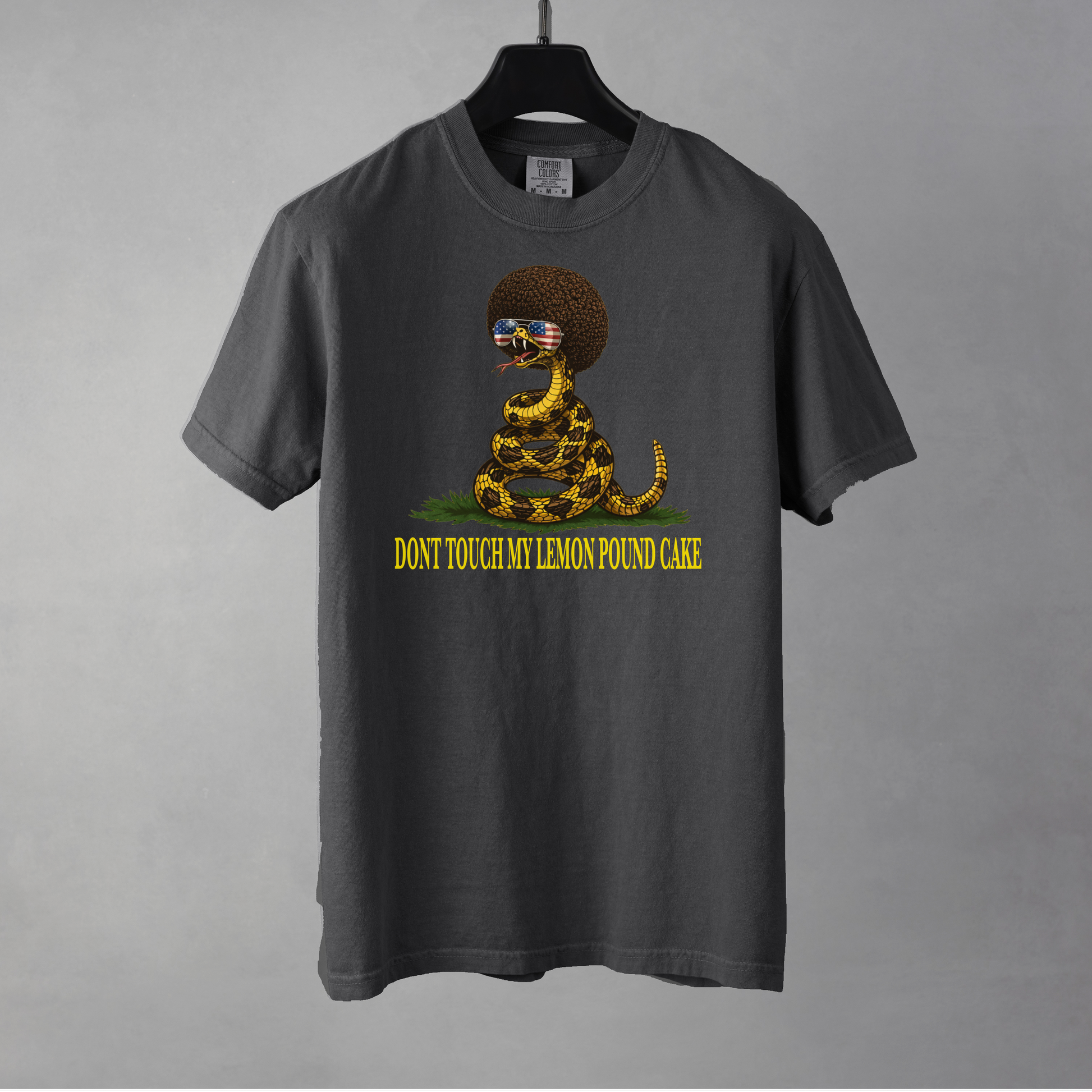 A pepper colored graphic t-shirt featuring a coiled rattlesnake with a large brown afro and flag-mirrored sunglasses above yellow text that reads "DONT TOUCH MY LEMON POUND CAKE." The shirt is resting on a black hanger against a gray background.