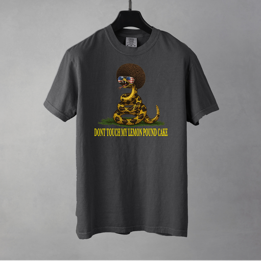A pepper colored graphic t-shirt featuring a coiled rattlesnake with a large brown afro and flag-mirrored sunglasses above yellow text that reads "DONT TOUCH MY LEMON POUND CAKE." The shirt is resting on a black hanger against a gray background.