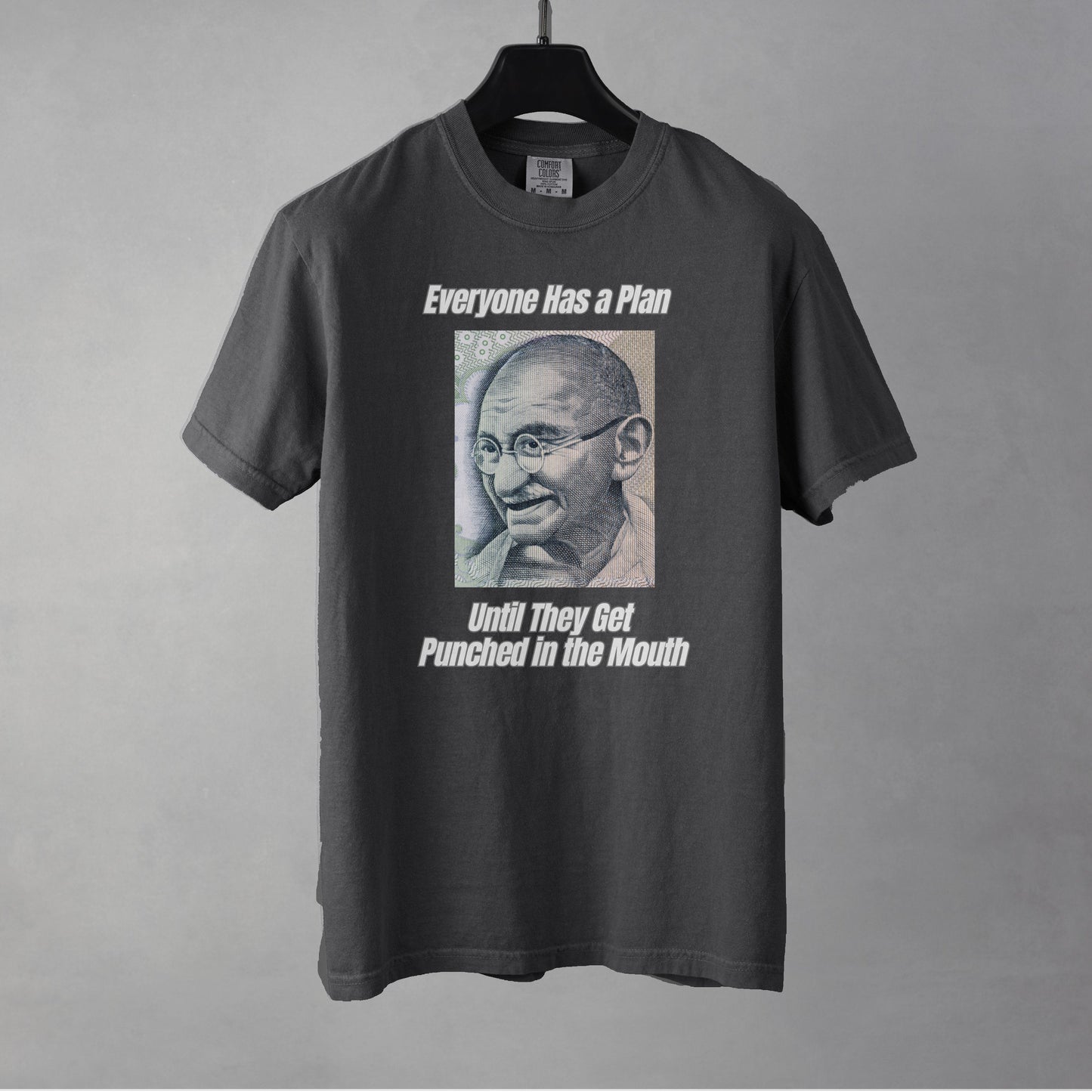 A pepper colored women's graphic t-shirt featuring a vintage portrait of Mahatma Gandhi ironically paired with the famous quote 'Everyone Has a Plan Until They Get Punched in the Mouth'