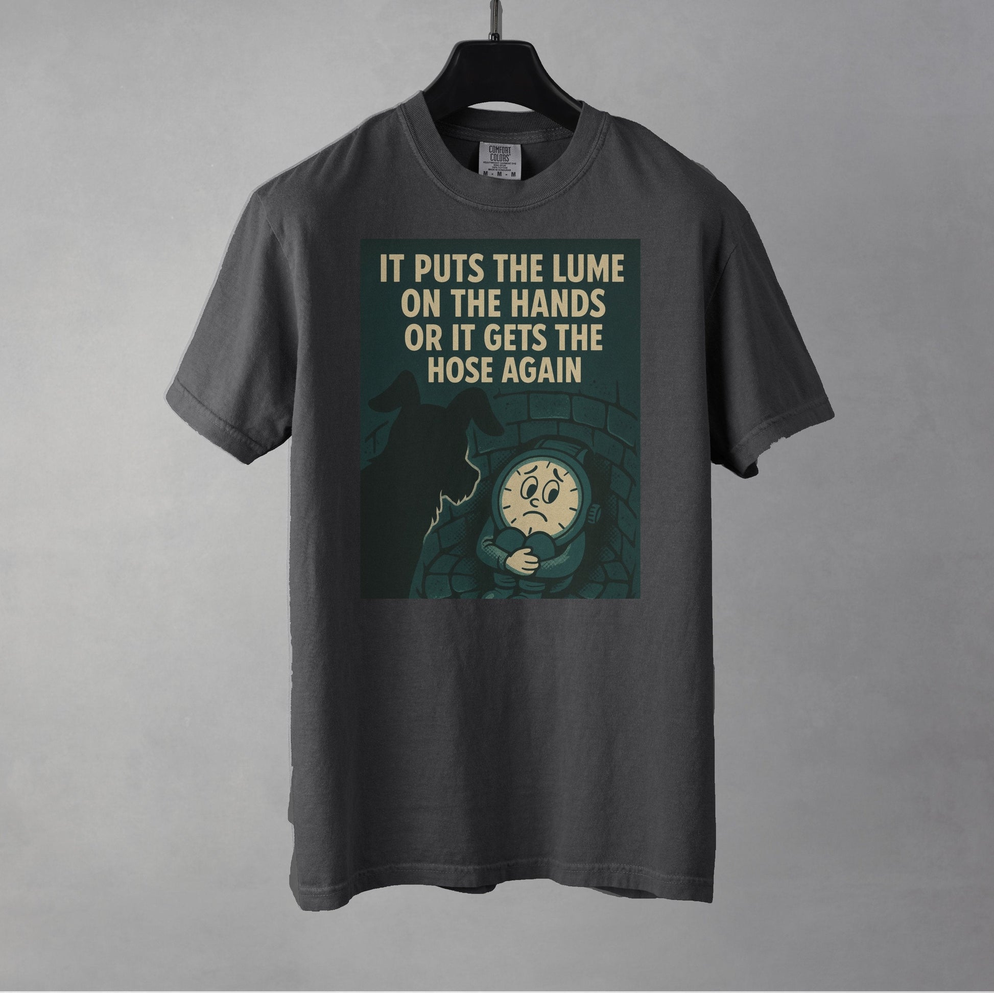 A pepper colored t-shirt on a black clothes hanger against a gray background. A graphic in dark grayish green with a cream colored line drawing of an anamorphic watch face sitting at the bottom of a brick lined well with its knees drawn up to its chest, held by its arms. Looking down into the well is the silhouette of a small dog. The top of the graphic shows the text, in all caps, 'IT PUTS THE LUME ON THE HANDS OR IT GETS THE HOSE AGAIN'.