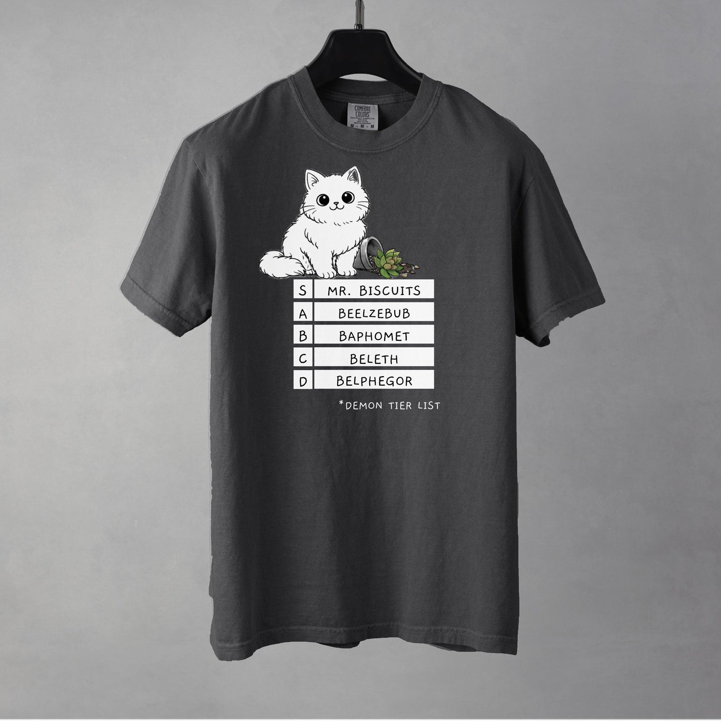A pepper colored t-shirt with a graphic of a white, fluffy kitten standing behind a knocked over plant. Beneath the cat is a 'Demon Tier List' with the cat 'Mr. Biscuits' listed atop the list in the S tier position.