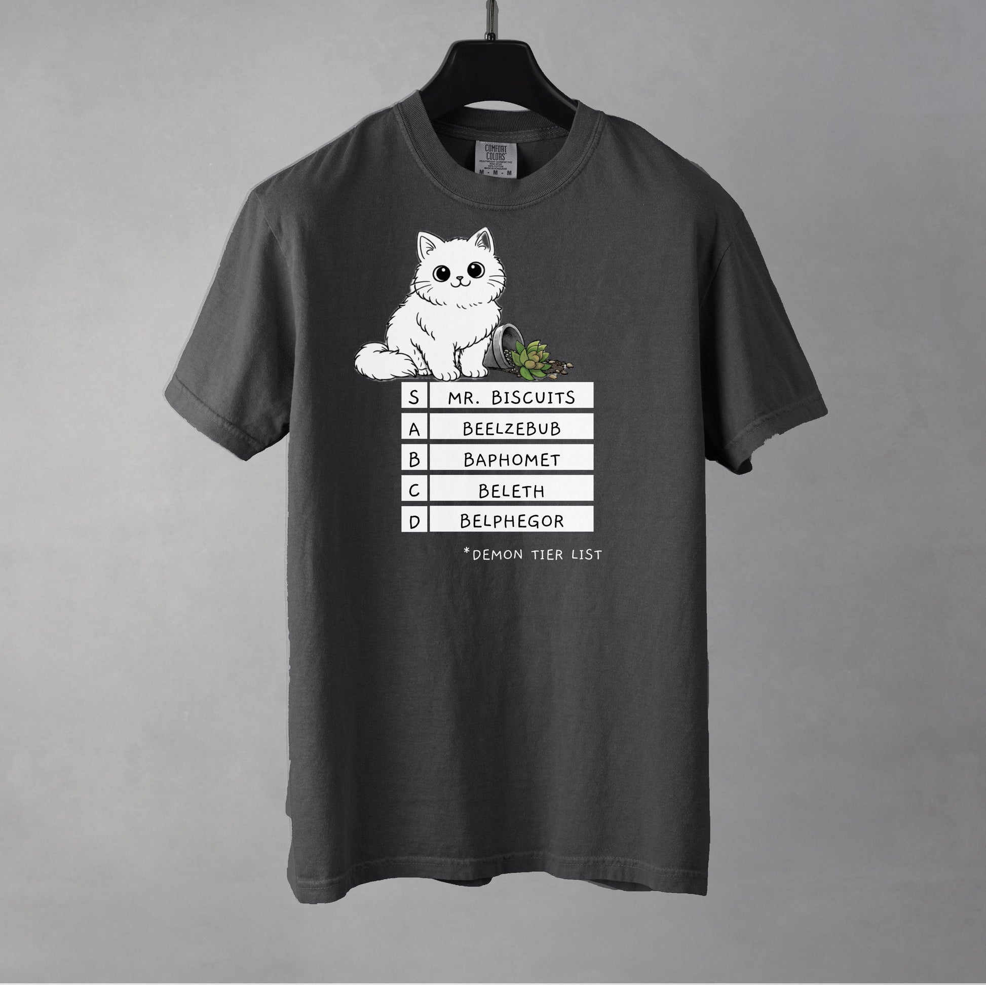 A pepper colored t-shirt with a graphic of a white, fluffy kitten standing behind a knocked over plant. Beneath the cat is a 'Demon Tier List' with the cat 'Mr. Biscuits' listed atop the list in the S tier position.
