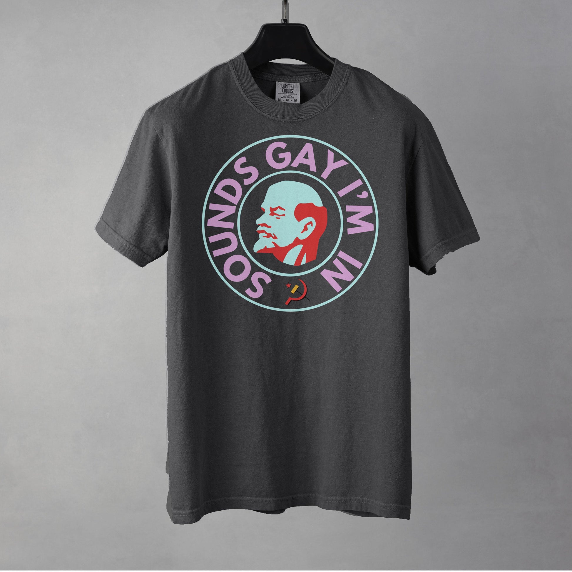 A pepper colored crew neck t-shirt hung from a black hanger against a gray background. The chest shows a graphic of an art deco style bust of Lenin's right profile with a light blue face and red hair, beard and eyebrows surrounded by a line circle in the same light blue. An outer circle of bold all caps neon pink text reads, 'SOUNDS GAY I'M IN' beginning at the 7 o'clock position. It is surrounded by the same light blue circle. 