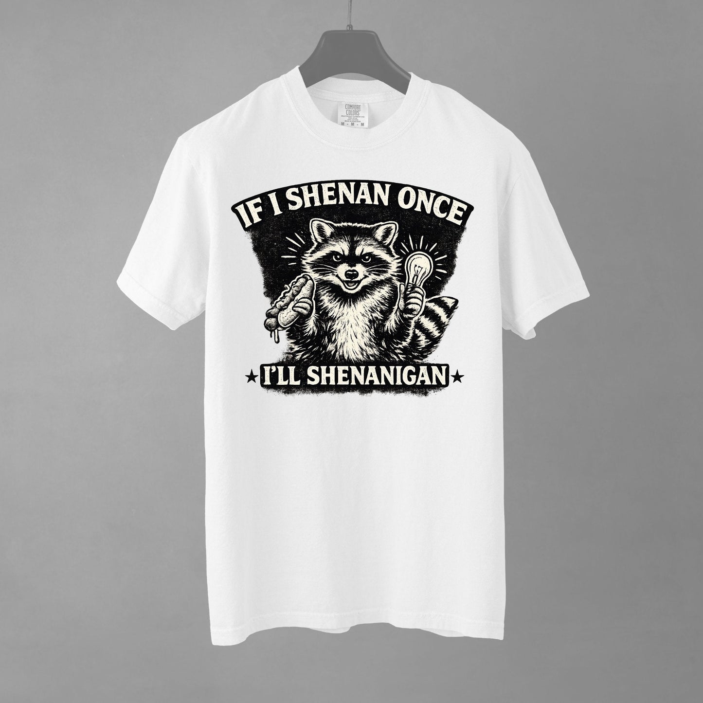 A white short-sleeve crewneck t-shirt featuring a distressed black and white illustration of a smiling raccoon holding a hot dog in one paw and a glowing lightbulb in the other. Arched above the raccoon is text that reads 'IF I SHENAN ONCE', and below the raccoon is text flanked by stars that reads 'I'LL SHENANIGAN'.