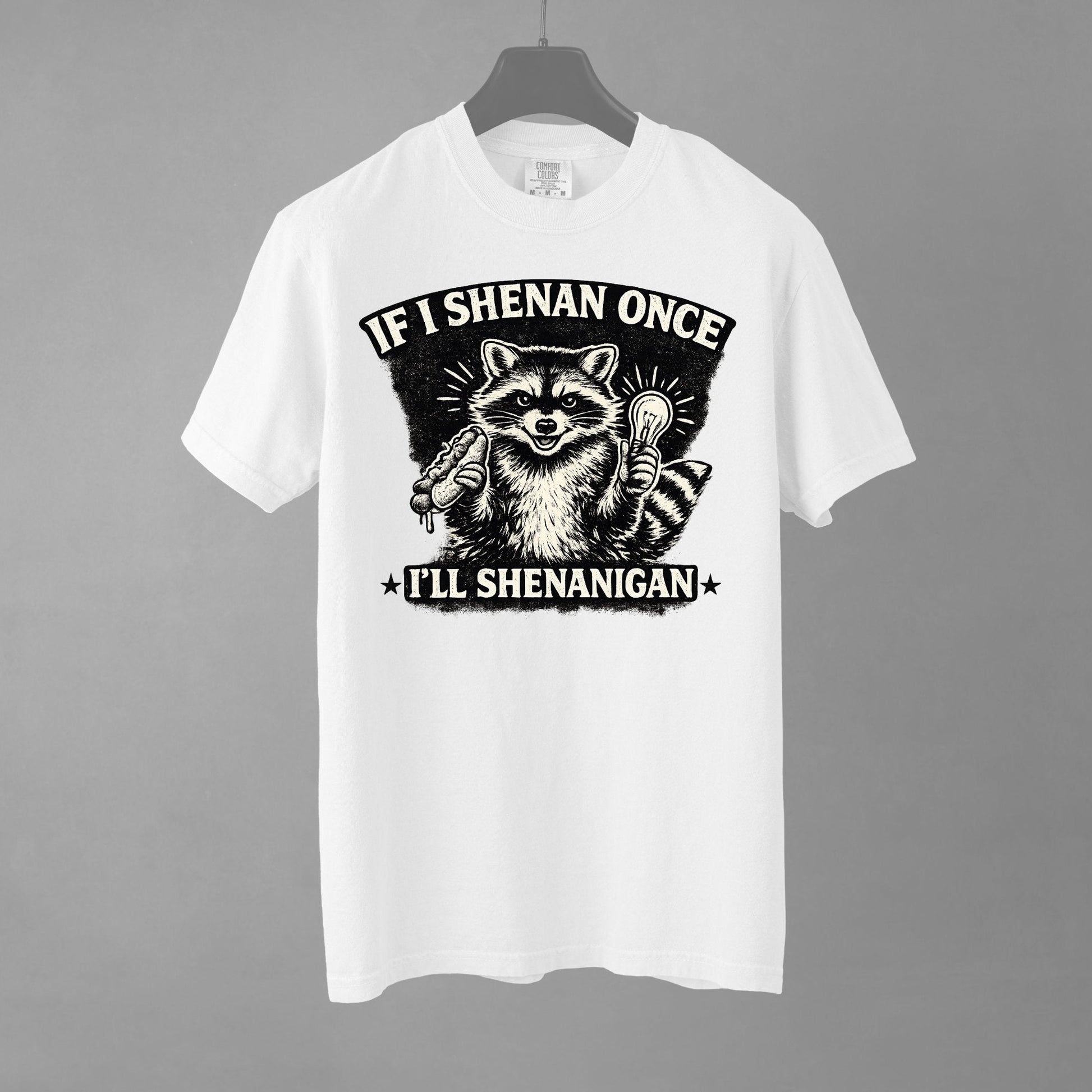 A white short-sleeve crewneck t-shirt featuring a distressed black and white illustration of a smiling raccoon holding a hot dog in one paw and a glowing lightbulb in the other. Arched above the raccoon is text that reads 'IF I SHENAN ONCE', and below the raccoon is text flanked by stars that reads 'I'LL SHENANIGAN'.