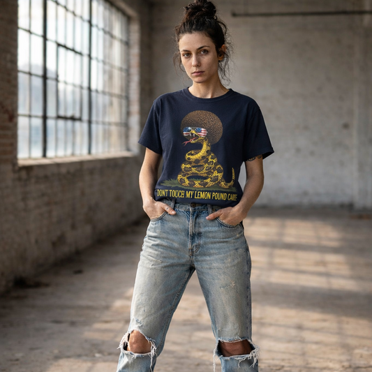 A woman standing defiantly with her hands in her pockets in an industrial loft, looking directly at the viewer. She is wearing a black graphic t-shirt featuring a coiled rattlesnake with a large brown afro and flag-mirrored sunglasses above yellow text that reads "DONT TOUCH MY LEMON POUND CAKE."